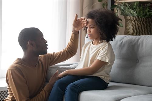 Young girl sitting on sofa with dad checking forehead for temperature
